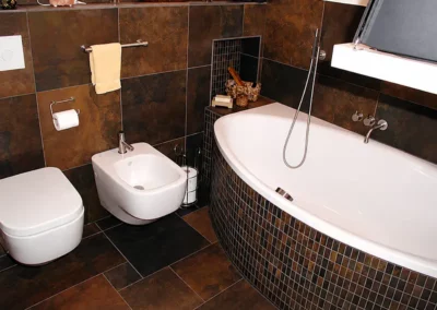 Modernes Badezimmer mit dunkelbraunen Wandfliesen und Bodenfliesen in Natursteinoptik, Eckbadewanne mit Mosaikfliesen sowie wandhängendem WC und Bidet.