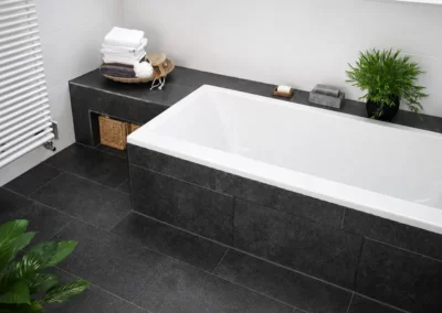 Modernes Badezimmer unter der Dachschräge mit weißer Badewanne, dunkelgrauen Bodenfliesen und Wandfliesen in Natursteinoptik.
