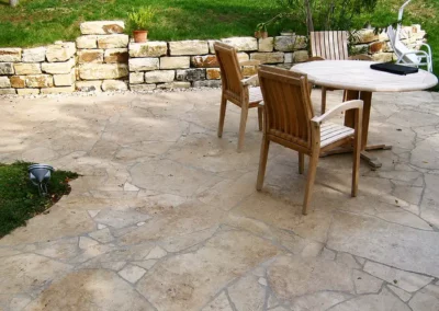 Terrasse mit unregelmäßig verlegten Natursteinplatten in Beige, rustikaler Natursteinmauer und Sitzbereich mit Holzgartenmöbeln.