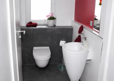 Modernes Gäste WC mit dunkelgrauen Bodenfliesen und Wandfliesen in Natursteinoptik, wandhängender Toilette, weißem Standwaschbecken und roter Akzentwand.
