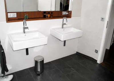 Modernes Badezimmer mit dunkelgrauen Bodenfliesen in Natursteinoptik, weißen Wandfliesen und zwei wandhängenden Waschbecken mit Spiegeln.