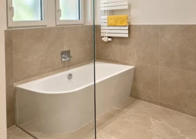 Modernes Badezimmer mit weißer Badewanne, beigen Bodenfliesen und Wandfliesen in Natursteinoptik, Glasabtrennung und weißem Handtuchheizkörper.