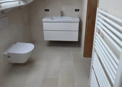 Modernes Dachgeschossbad mit hellbeigen Bodenfliesen und Wandfliesen in Natursteinoptik, wandhängendem WC, weißem Waschtisch und Handtuchheizkörper.