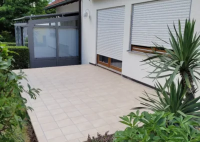 Geflieste Terrasse mit hellbeigen Terrassenfliesen, angrenzendem Wintergarten und grüner Gartenbepflanzung.