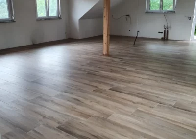 Großzügiger Wohnraum im Dachgeschoss mit hellbraunen Bodenfliesen in Holzoptik, natürlicher Holzstütze und modernem Fliesenbelag.